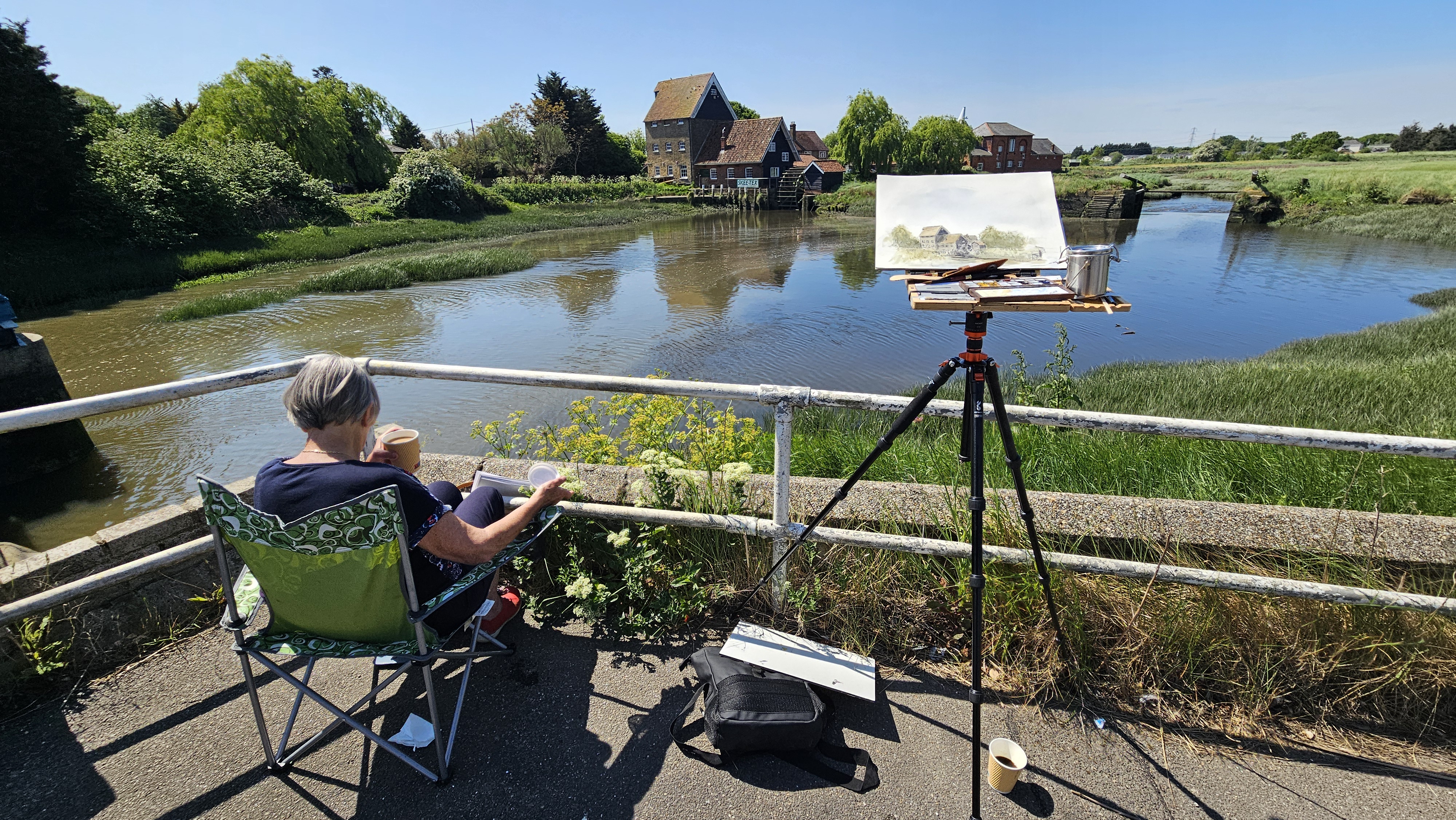 Painting a watercolour of a riverside mill at Battlesbridge, Essex, easel set up beside the water with the artist seated and sketching