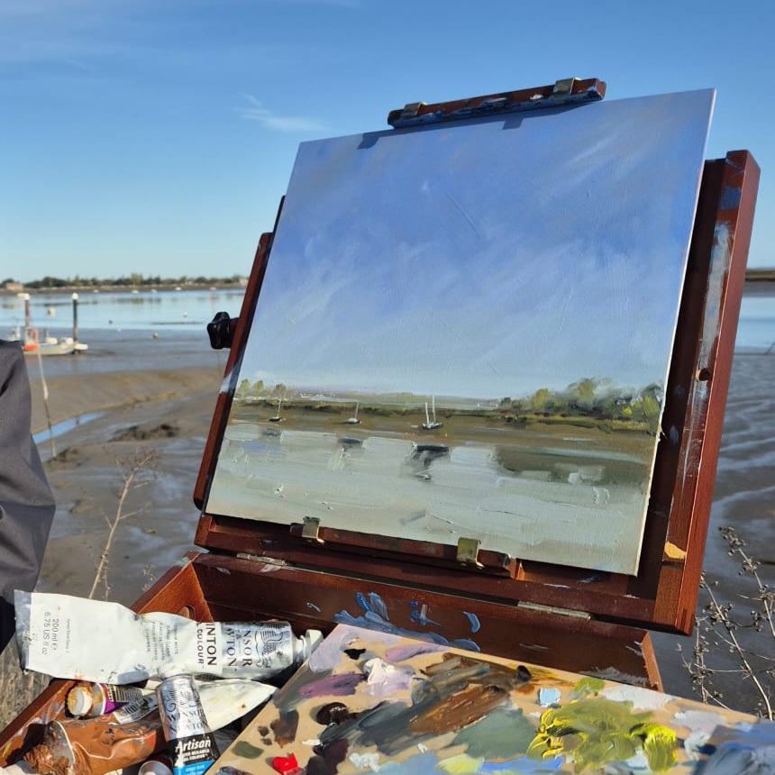 Painting set up on an easel at low tide at Heybridge Basin near Maldon, Essex, working on a river scene with moored boats